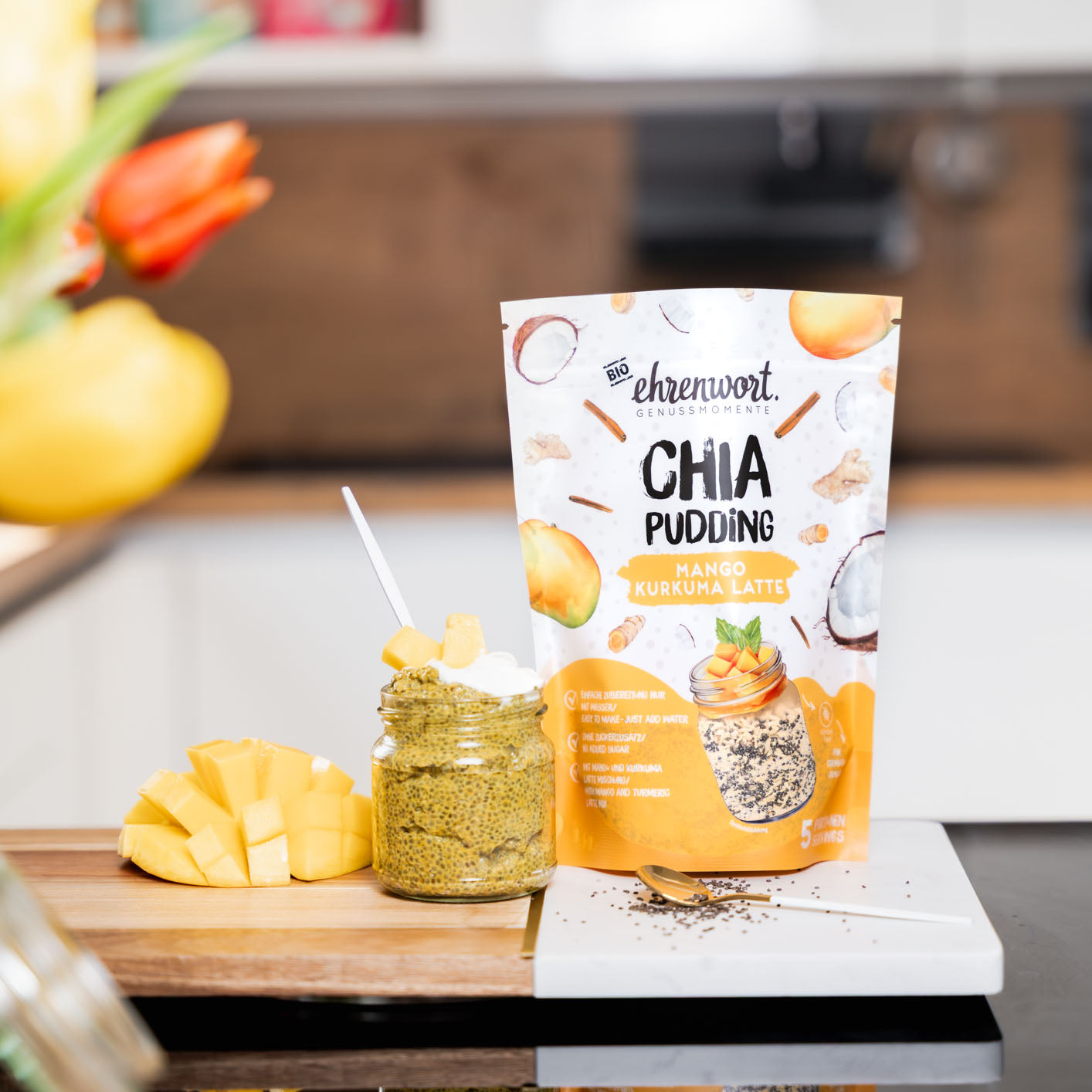 ehrenwort BIO Mango Kurkuma Latte Chia Pudding Verpackung neben zubereiteter Portion in einer Küche auf einem Schneidebrett